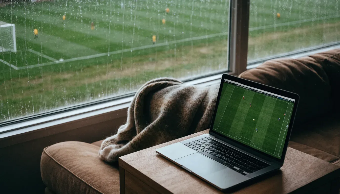 Ein Sofa mit Wolldecke steht vor einem grossen Fenster, draussen Regen und in der Ferne ein Fussballplatz.