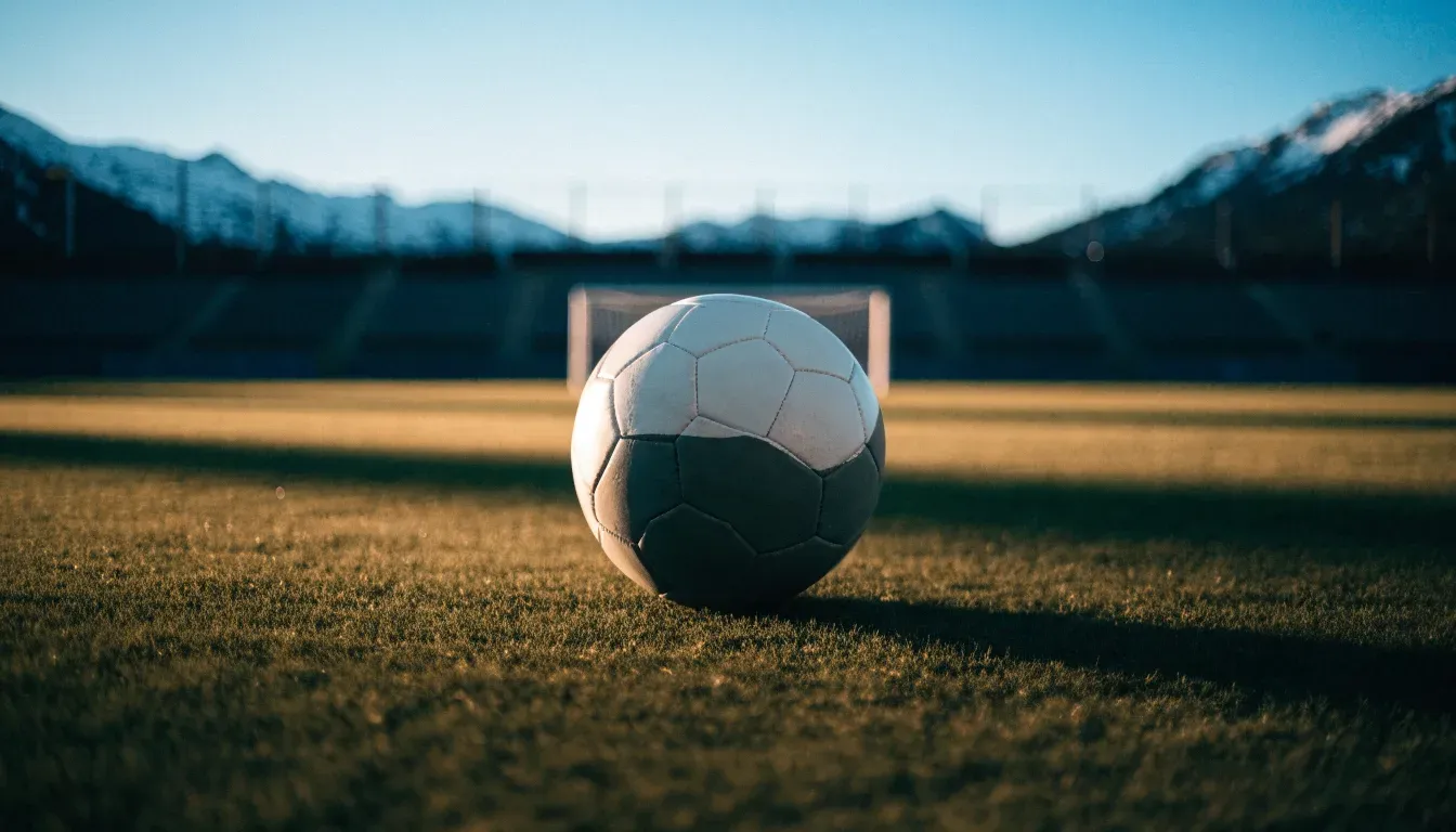 Ein Fussball liegt auf dem Rasen eines leeren Stadions am frühen Abend, dahinter erheben sich schneebedeckte Berge unter klarem Himmel.