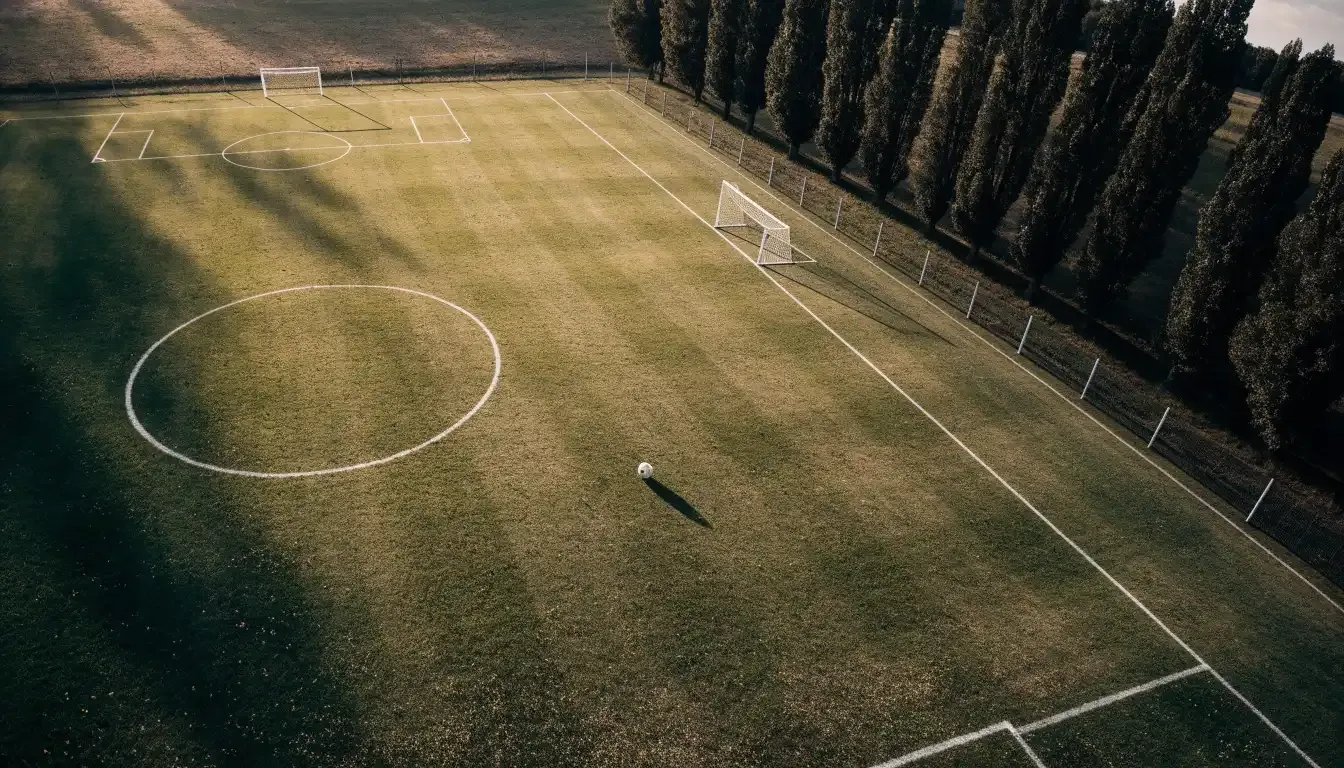 Ein leerer Fussballplatz mit weissen Linien von oben gesehen im Morgenlicht, am Rand stehen zwei Tore und eine Reihe Pappeln.