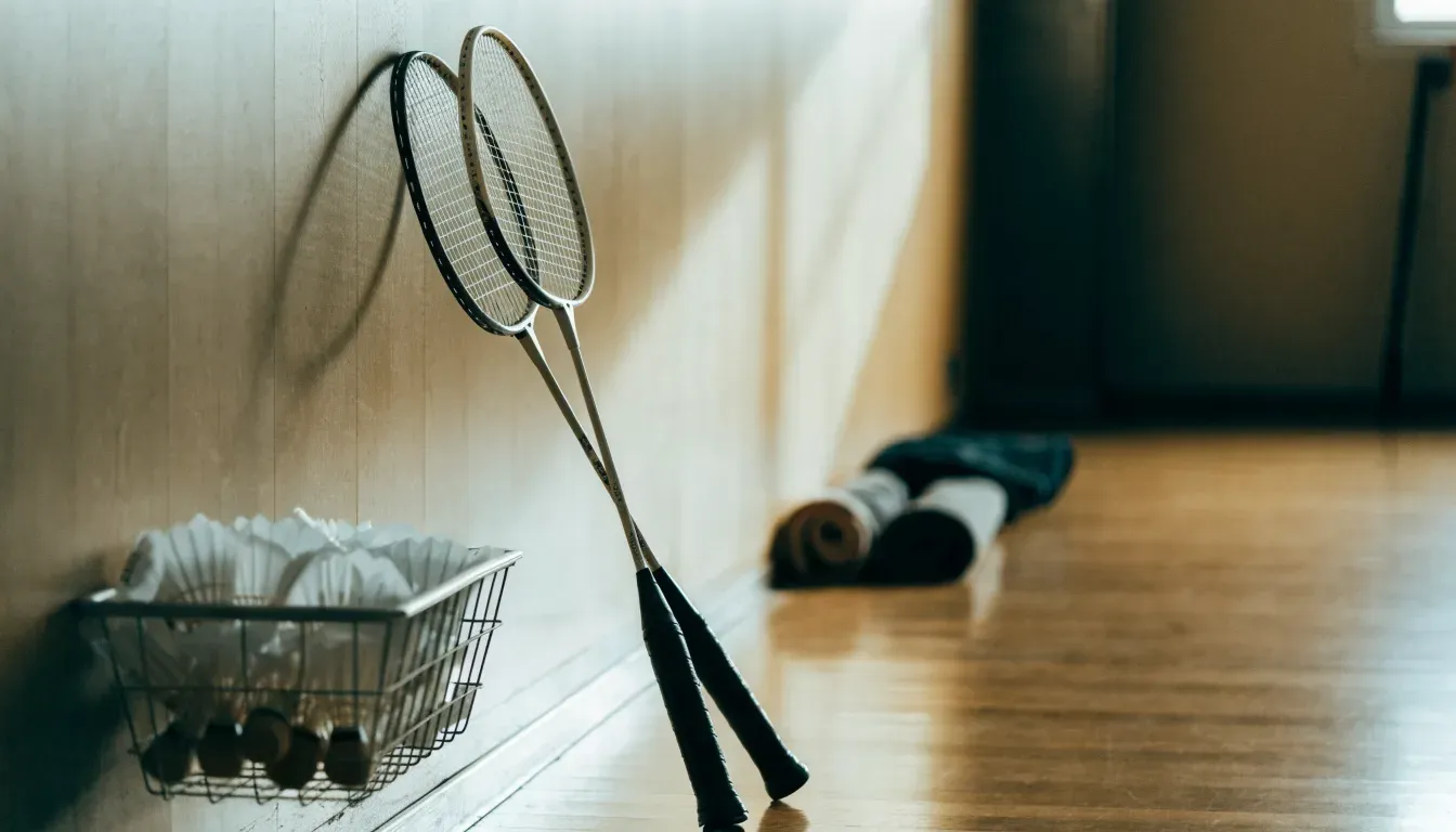 Zwei Badmintonschläger lehnen gekreuzt an einer Hallenwand aus Holz, daneben steht ein Korb mit Federbällen.