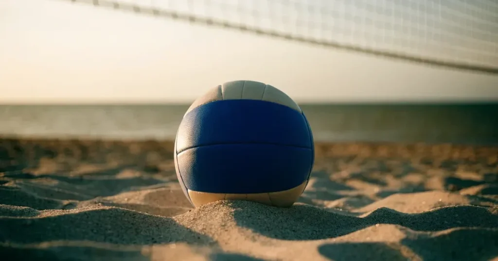 Ein Volleyball liegt auf feinem hellem Sand, ein gespanntes Netz verschwimmt dahinter, das Licht kommt von einem weiten Meer.