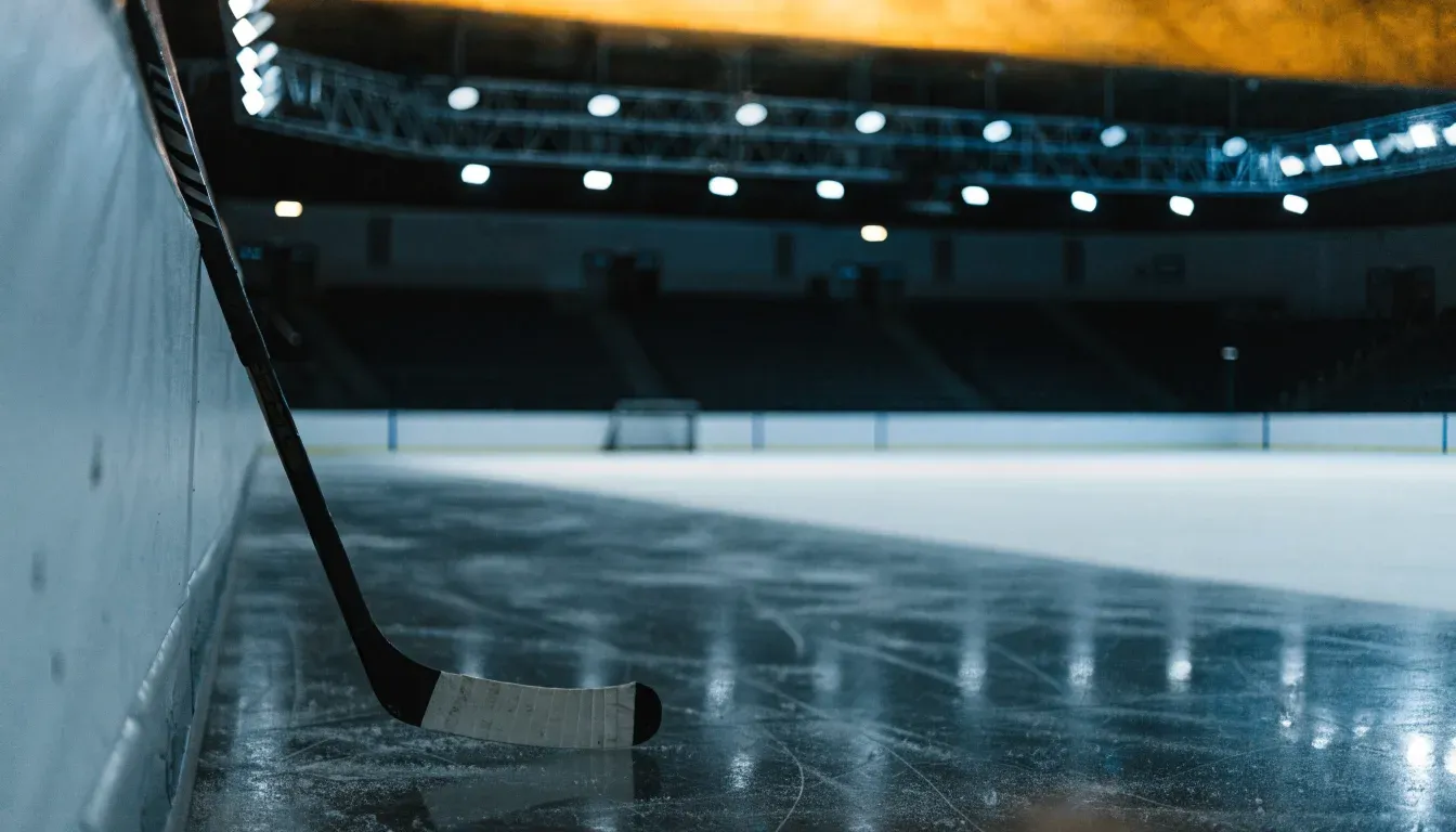 Ein Eishockeyschläger lehnt an der Bande, in der Ferne liegt die hell beleuchtete Eisfläche einer Halle unter Flutlicht.