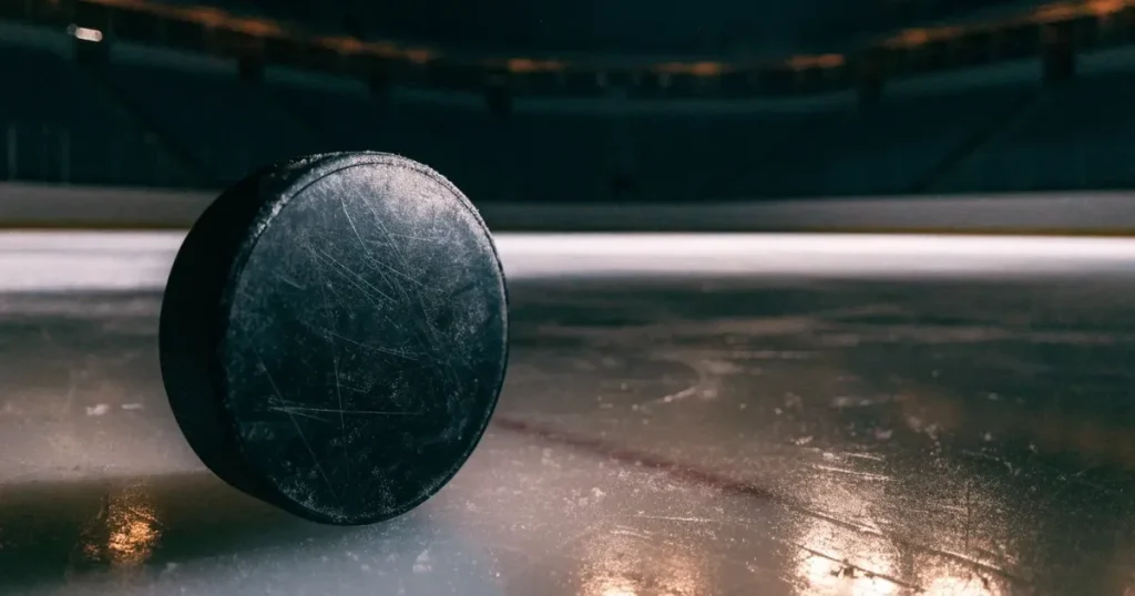Ein Eishockeypuck liegt auf einer glatten Eisfläche, im Hintergrund erheben sich die dunklen Ränge einer leeren Halle.
