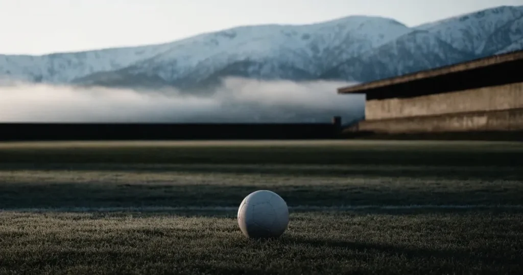 Ein Fussball liegt am frühen Morgen auf dem Rasen eines leeren Fussballfeldes, dahinter eine kleine Tribüne und schneebedeckte Berge.